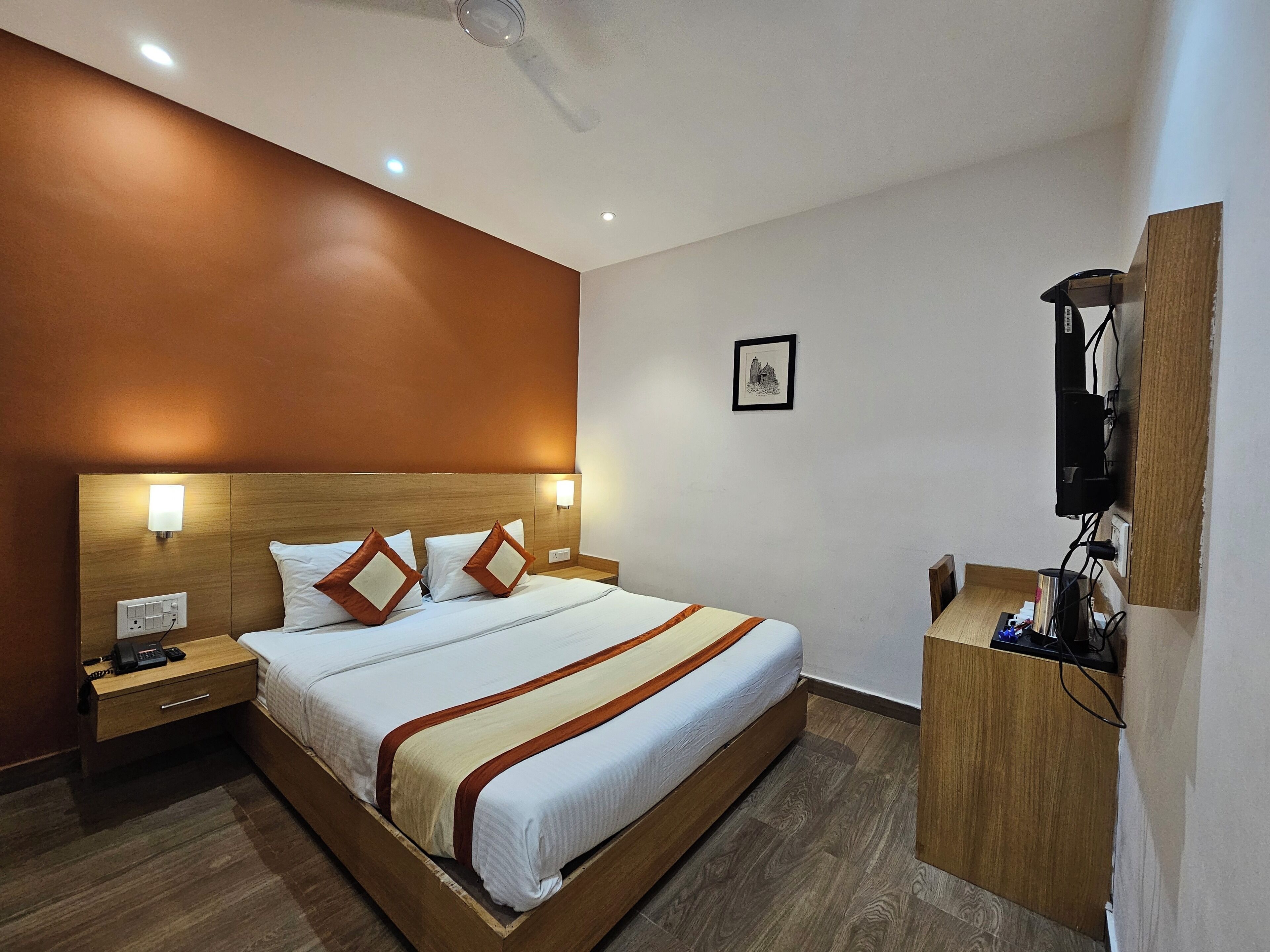 A hotel room with a wooden bed, a nightstand, a desk, and a wall-mounted TV.