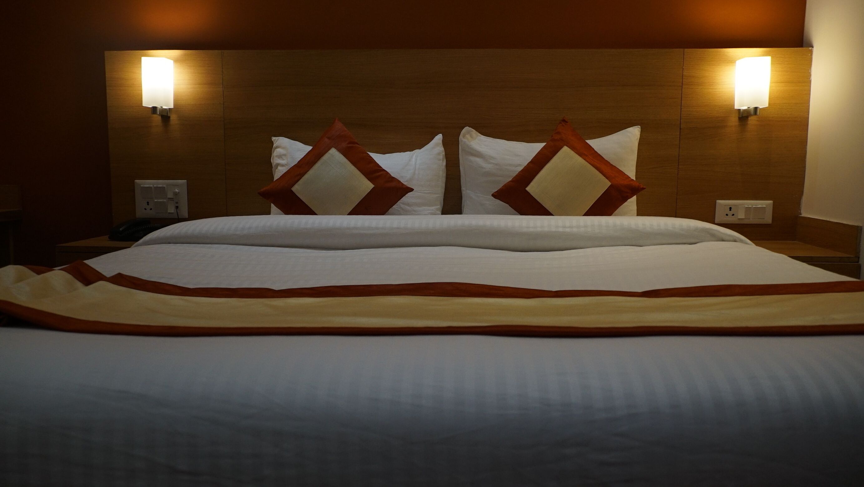 Een net opgemaakt bed met een wit-beige sprei, twee decoratieve kussens en twee wandlampen.
