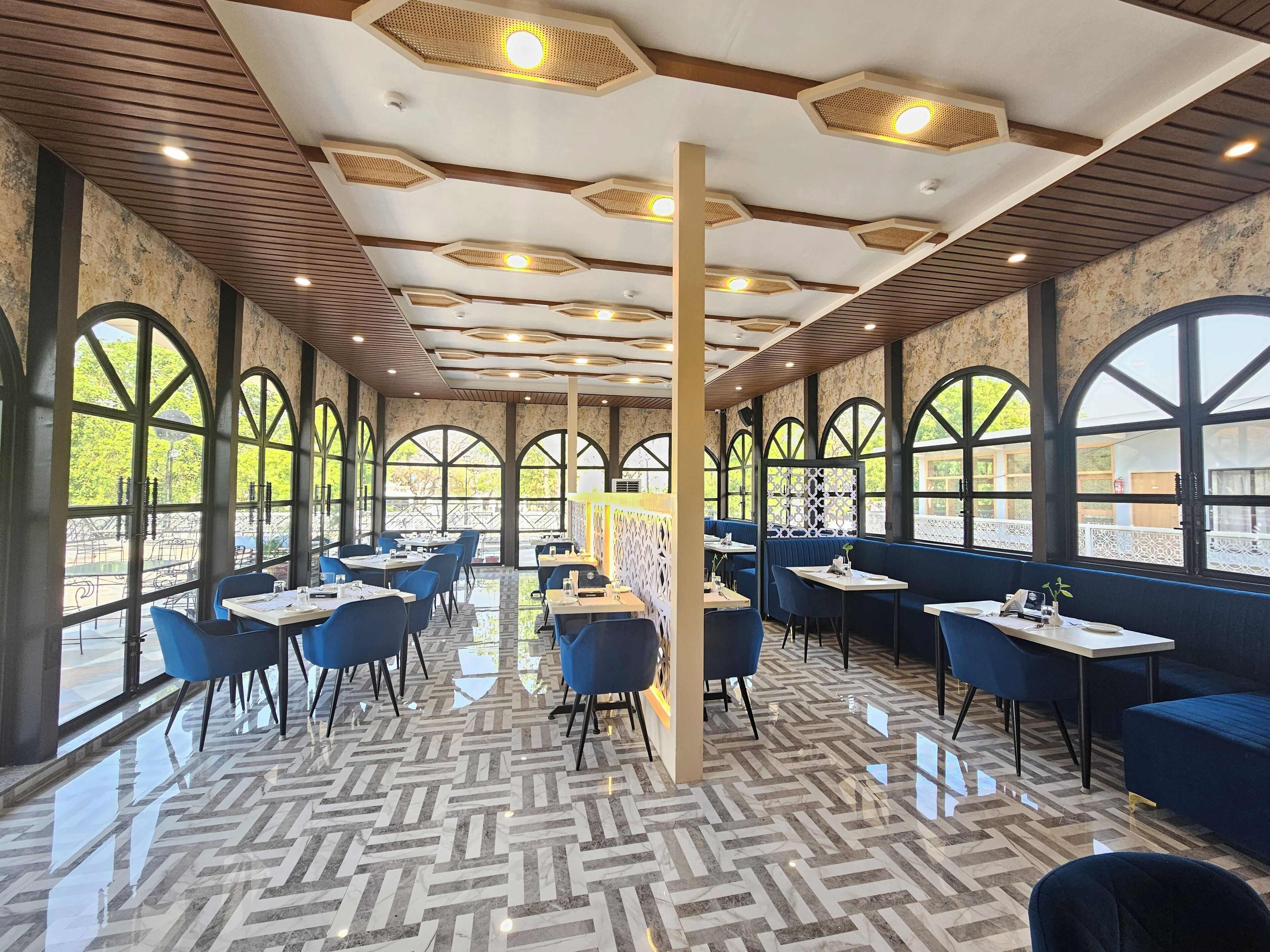 Un restaurante con una decoración moderna, que presenta luces de techo hexagonales, amplias ventanas en arco y un suelo de parqué pulido.