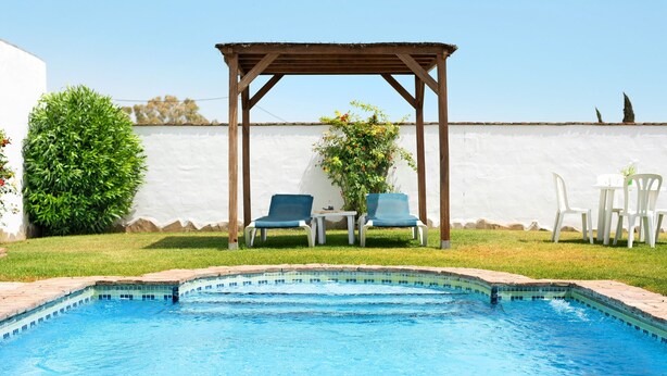 Piscina com pergolado de madeira, duas cadeiras e uma mesa.