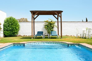 A swimming pool with a wooden pergola, two chairs, and a table.