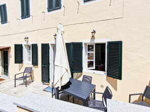 An outdoor seating area with tables and chairs, a covered umbrella, and a building with green shutters and a tiled floor.