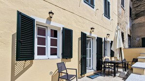 A building with multiple windows, some with shutters, a chair, a table, and an umbrella.