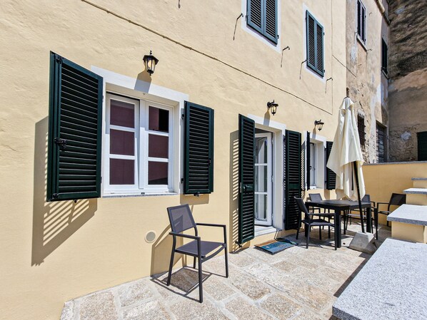 A building with multiple windows, some with shutters, a chair, a table, and an umbrella.