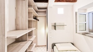 A bedroom with a bed, a wardrobe with multiple shelves, a window, and a tiled floor.