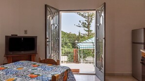 Una puerta abierta que da a un balcón con una reja de metal, un televisor sobre un mueble bajo, una mesa con mantel estampado y un refrigerador.