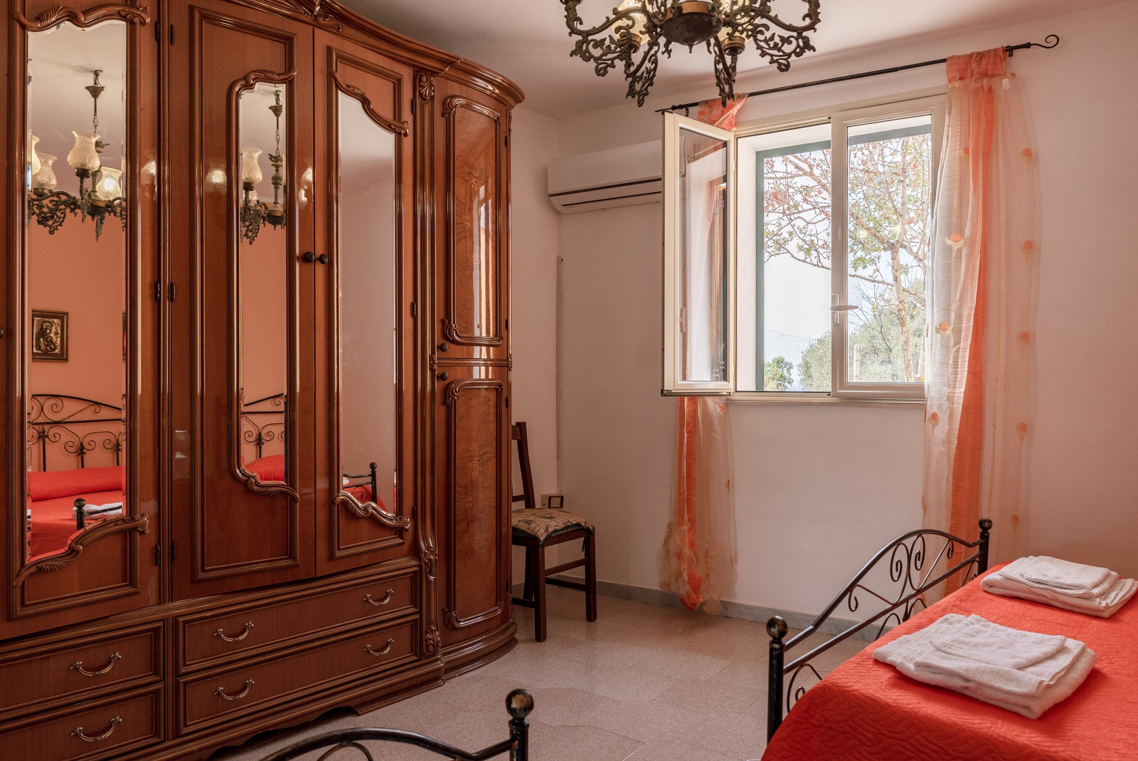 A bedroom with a wooden wardrobe, a chair, a bed with a red bedspread, and a window with curtains.