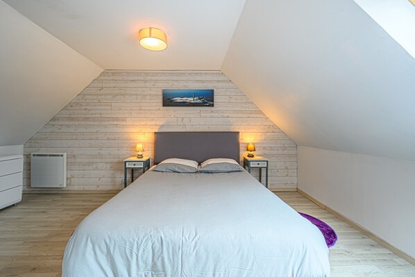 A bedroom with a sloped ceiling, a bed, two bedside lamps, a wall-mounted picture, and a window.