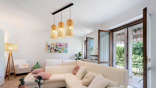 Un salon avec un canapé d’angle blanc, une petite table en bois, deux cactus en pot, un tableau accroché au mur et une grande fenêtre donnant sur un paysage verdoyant.