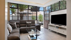 Een moderne woonkamer met een hoekbank, een glazen salontafel, een televisie op een witte dressoir en grote ramen met uitzicht op groen.