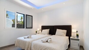 Hotelkamer met twee bedden, een nachtkastje met een lamp, een kleine potplant en uitzicht op palmbomen door een raam.