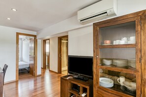 A modern living room with a flat-screen TV on a wooden stand, a glass-fronted cabinet with dishes, and a hallway leading to a bedroom.