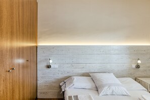 A room with a wooden wardrobe, a bed with white bedding, and two wall-mounted light fixtures.
