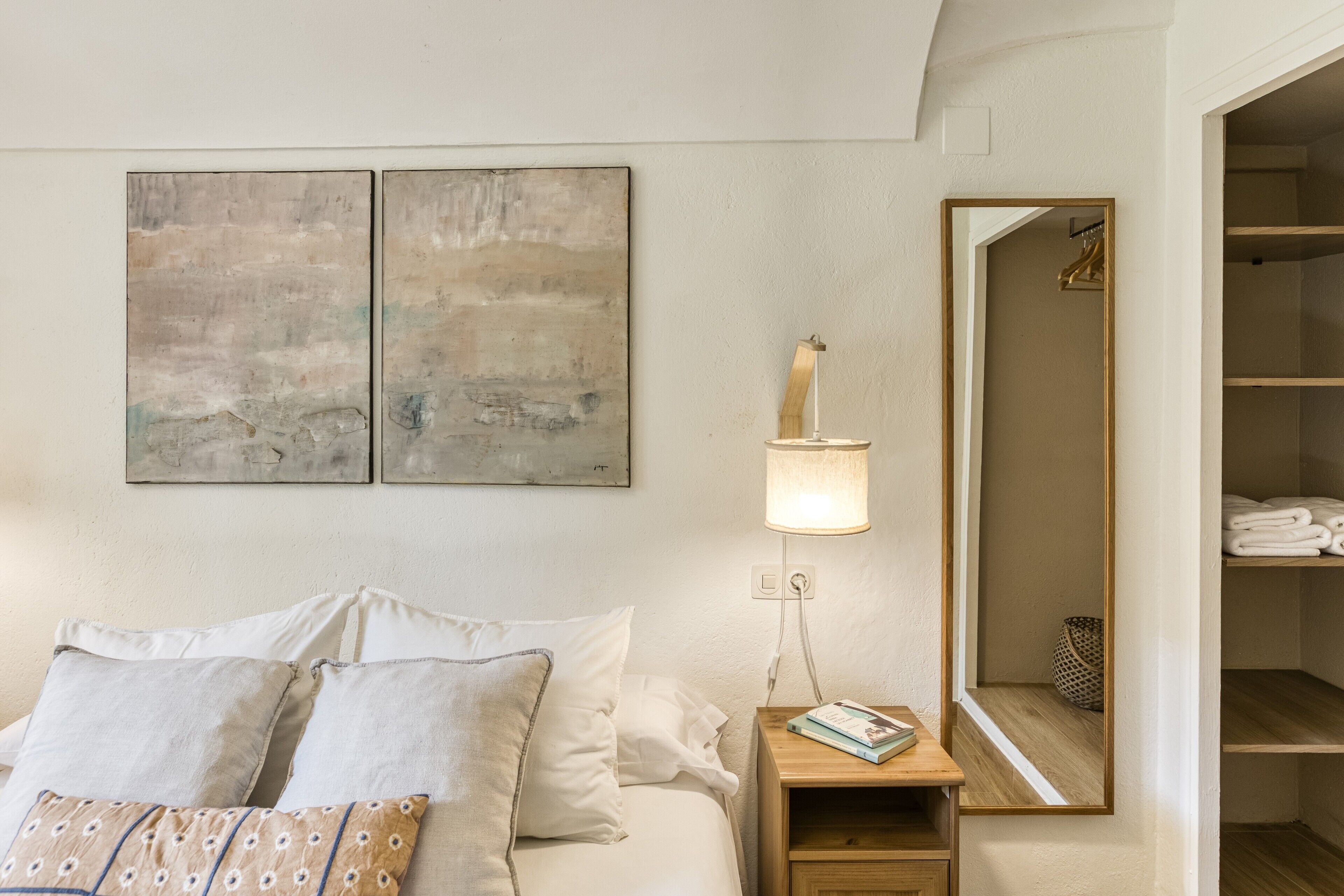 Ein ordentlich eingerichtetes Schlafzimmer mit einem Bett, Kissen, einem Nachttisch mit Lampe, einem Spiegel und einem Regal mit gefalteten Handtüchern.