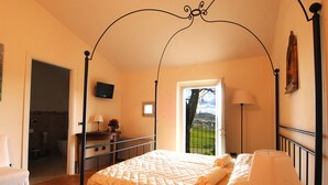Een slaapkamer met een hemelbed, een nachtkastje met een lamp, en uitzicht op een groen landschap. Een badkamer is zichtbaar door een geopende deur.