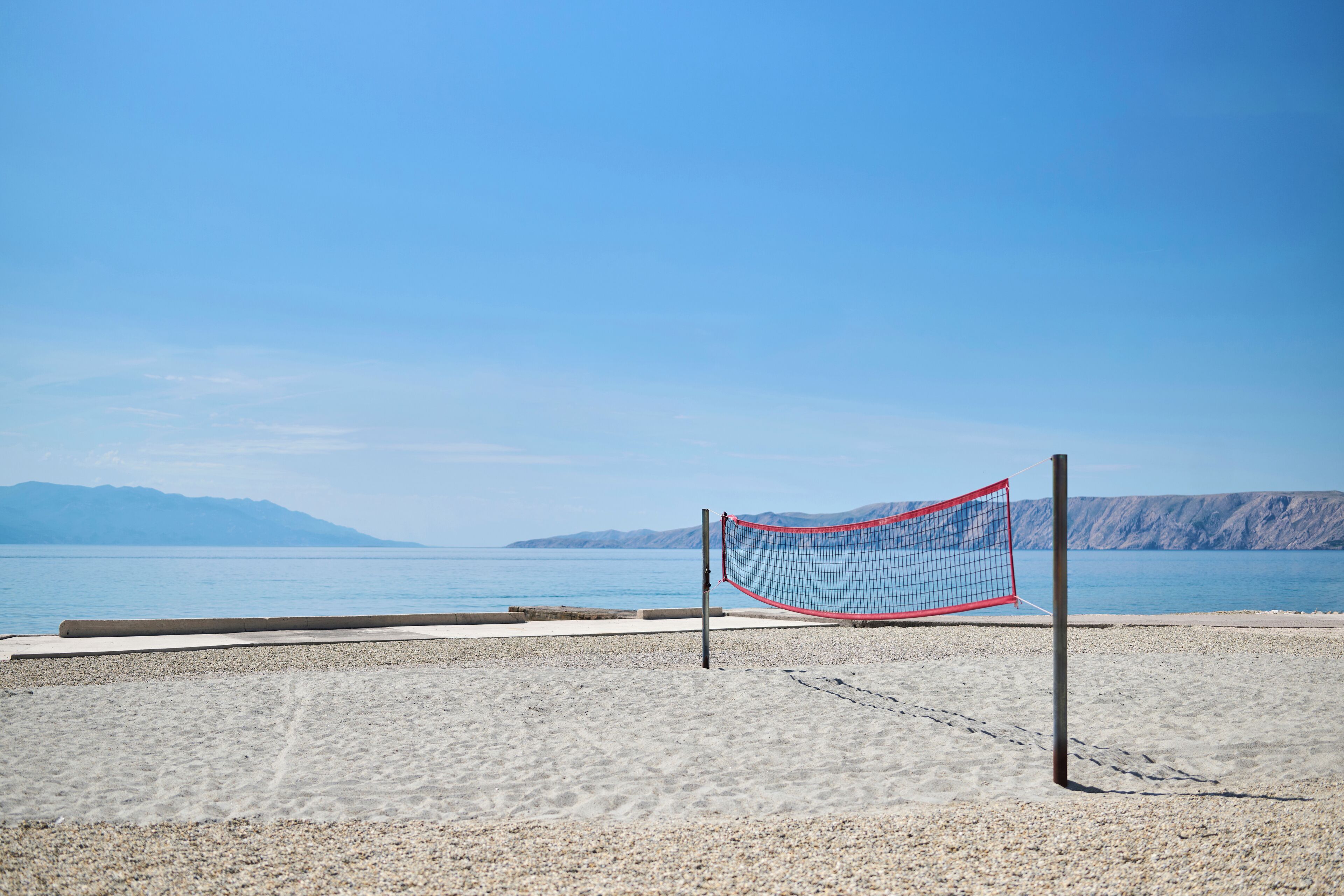 A beach volleyball net on a pebbled beach with a clear view of the sea and distant mountains.