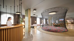 Un moderno vestíbulo de hotel con un mostrador de recepción de madera, una zona de estar curva con un tapete colorido y estampado, y una pantalla grande mostrando contenido.