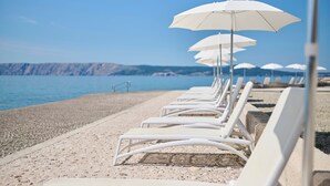 Una hilera de sillas reclinables blancas con sombrillas a lo largo de una playa de guijarros, frente a un mar tranquilo y colinas lejanas.