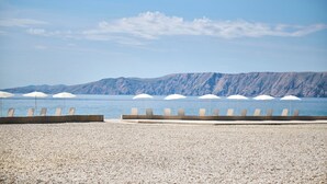 Una playa de guijarros con sombrillas blancas y sillones de descanso frente a un mar tranquilo y montañas.