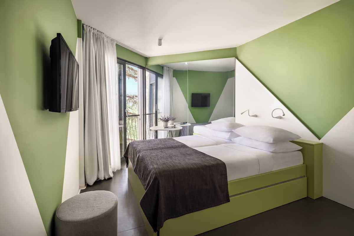 A modern hotel room with a bed, a TV, a small table, and a window with curtains.