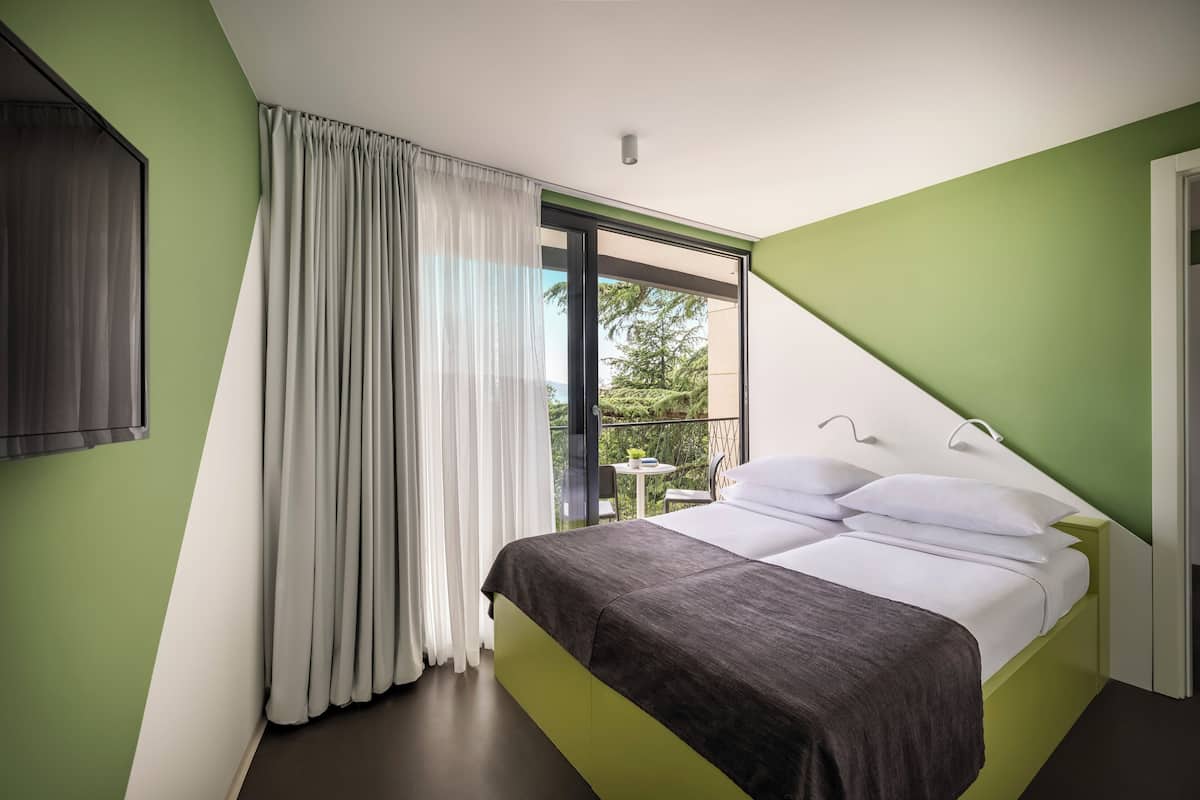 A modern bedroom with a bed, curtains, and a view of greenery outside.