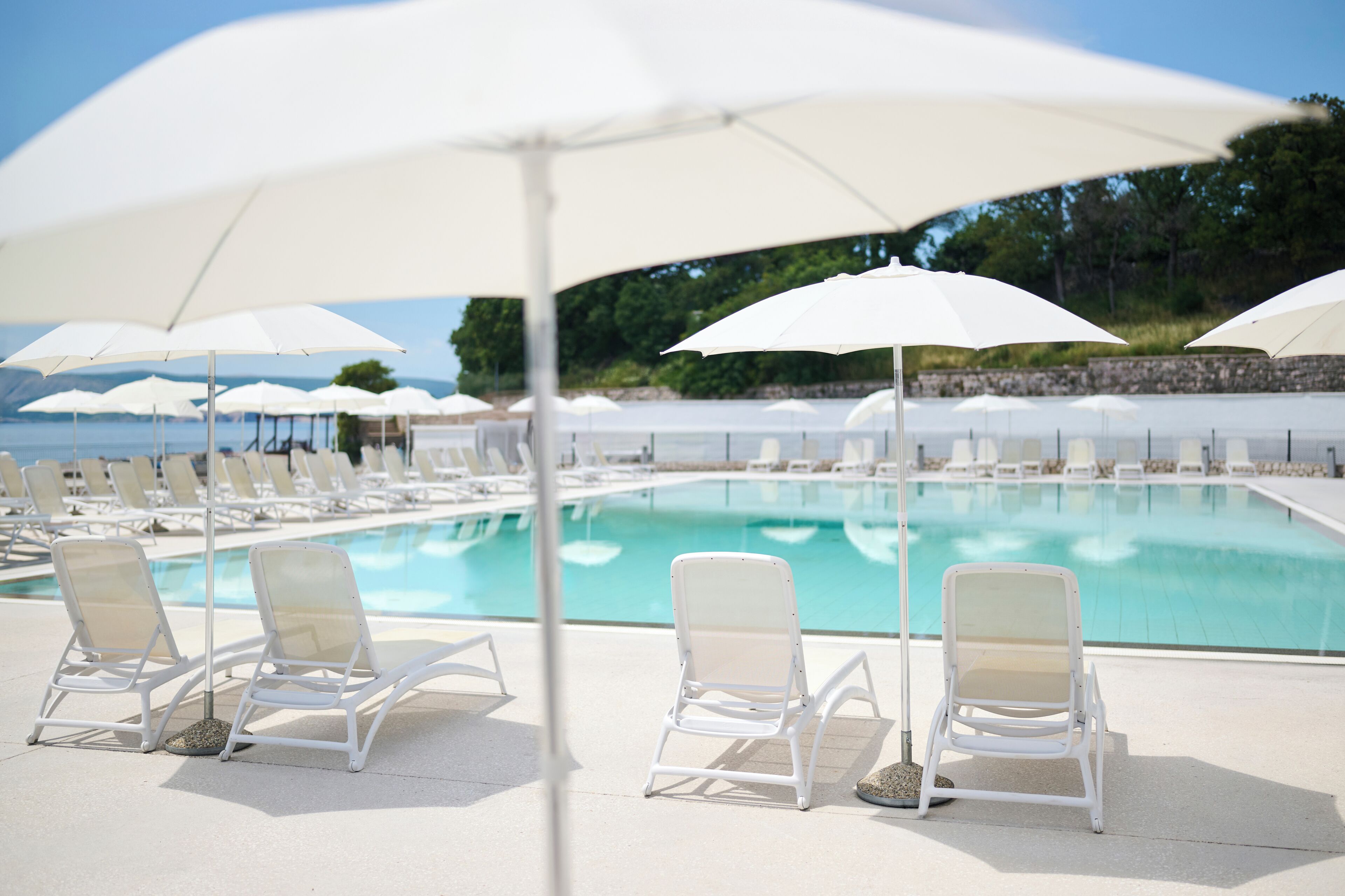 A poolside area with white lounge chairs and umbrellas, a clear pool, and a stone wall in the background.