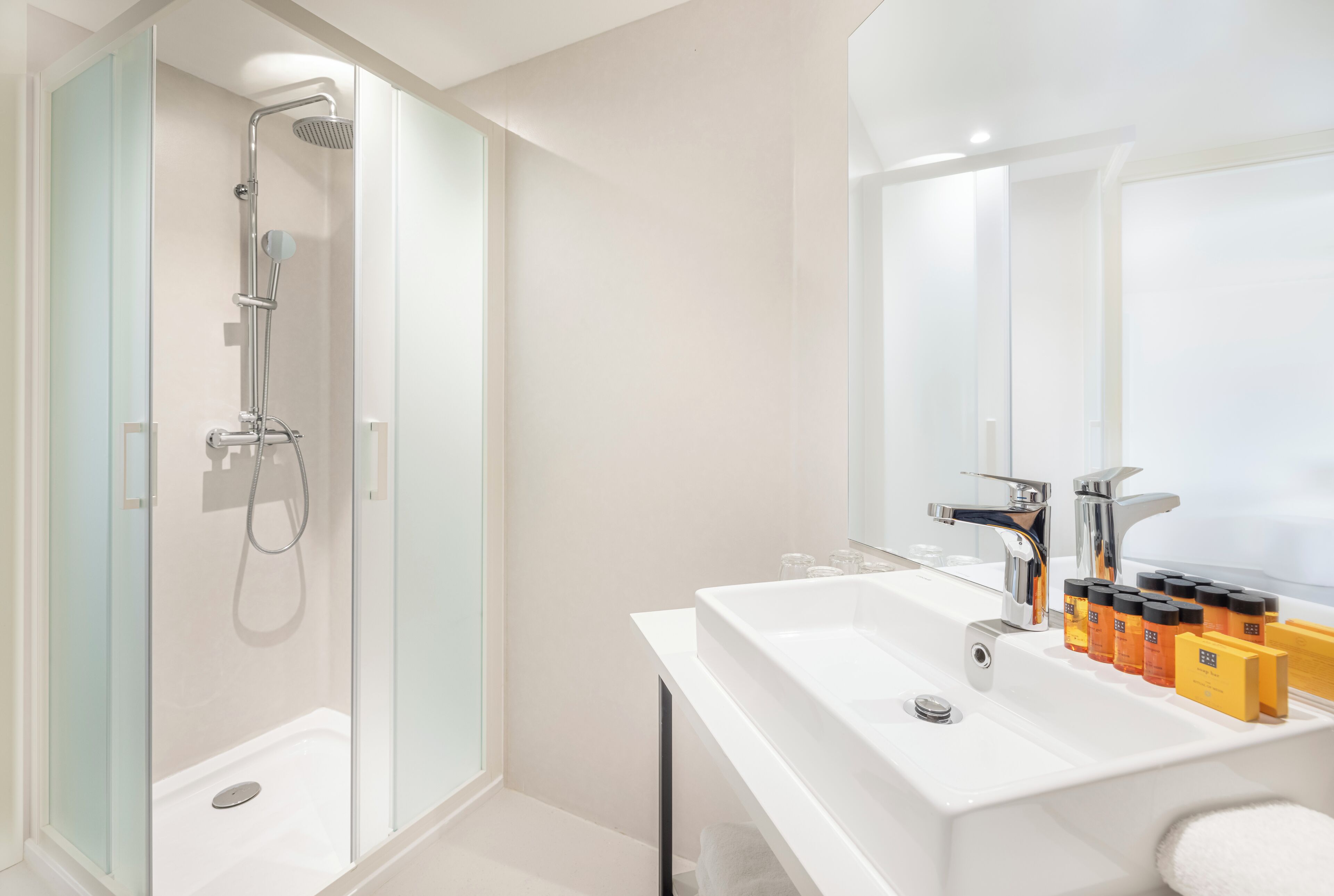 Un baño moderno con ducha de vidrio, lavamanos blanco con artículos de aseo y un espejo grande.