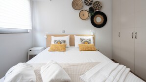 A neatly made bed with white bedding and patterned pillows, a bedside table with a lamp, and a wall decorated with woven wall hangings. - The Luxury Loft by Cloudkeys (Athens)