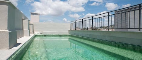 A rooftop swimming pool with clear blue water, surrounded by a white building and a metal railing.