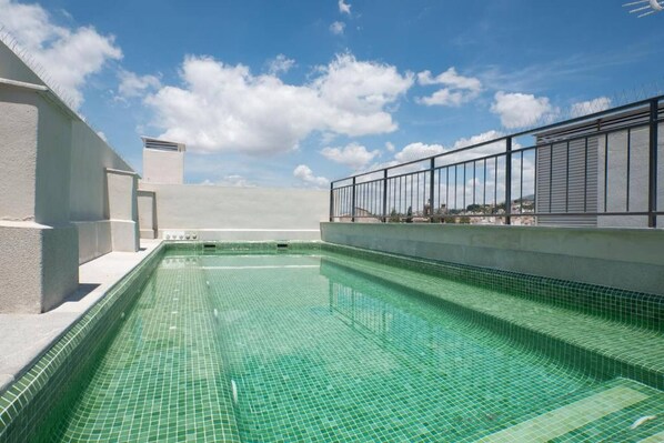 Una piscina sul tetto con acqua cristallina e azzurra, circondata da un edificio bianco e una ringhiera metallica.