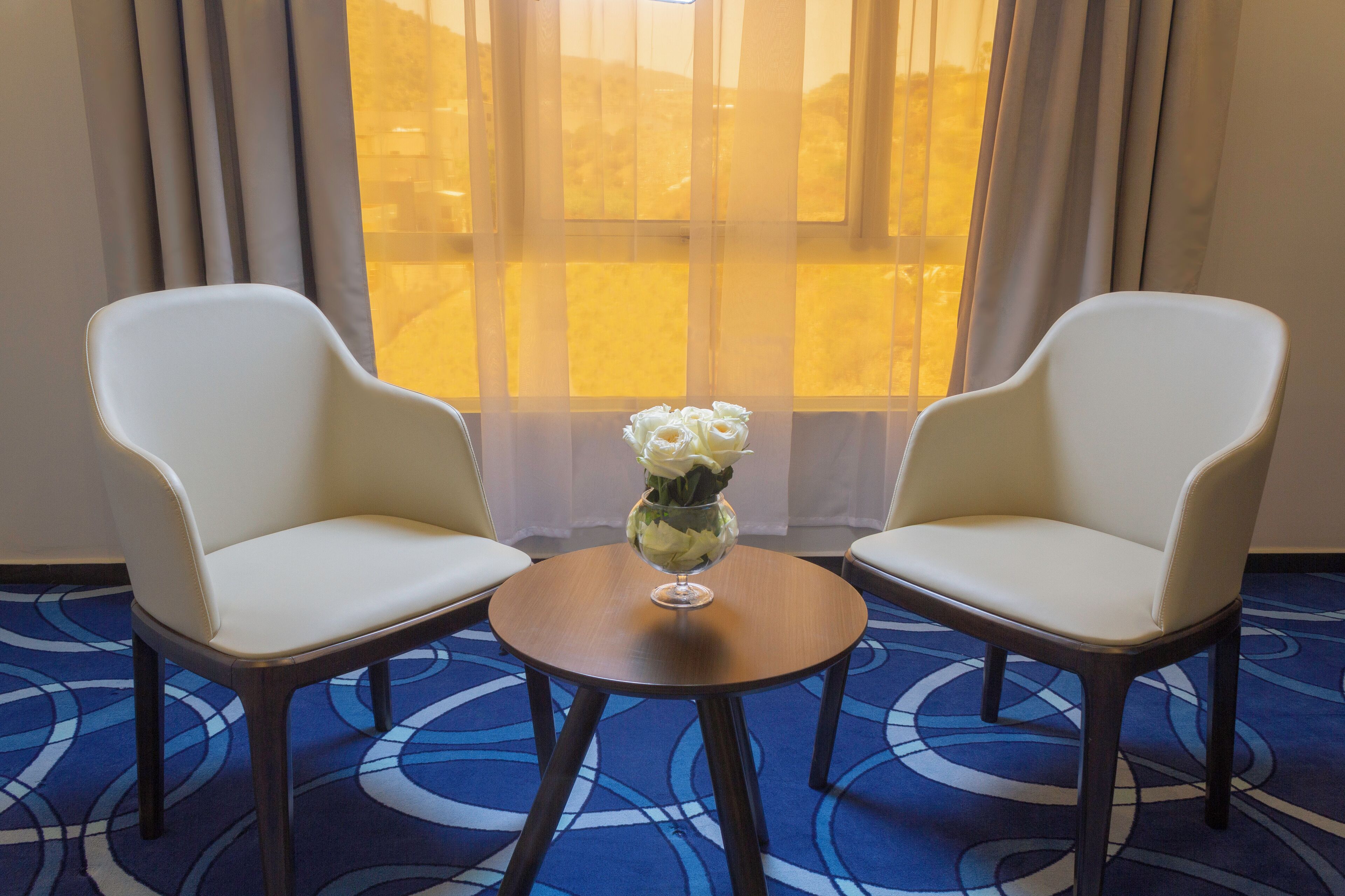 A room with two white chairs, a small round table, and a vase with flowers.