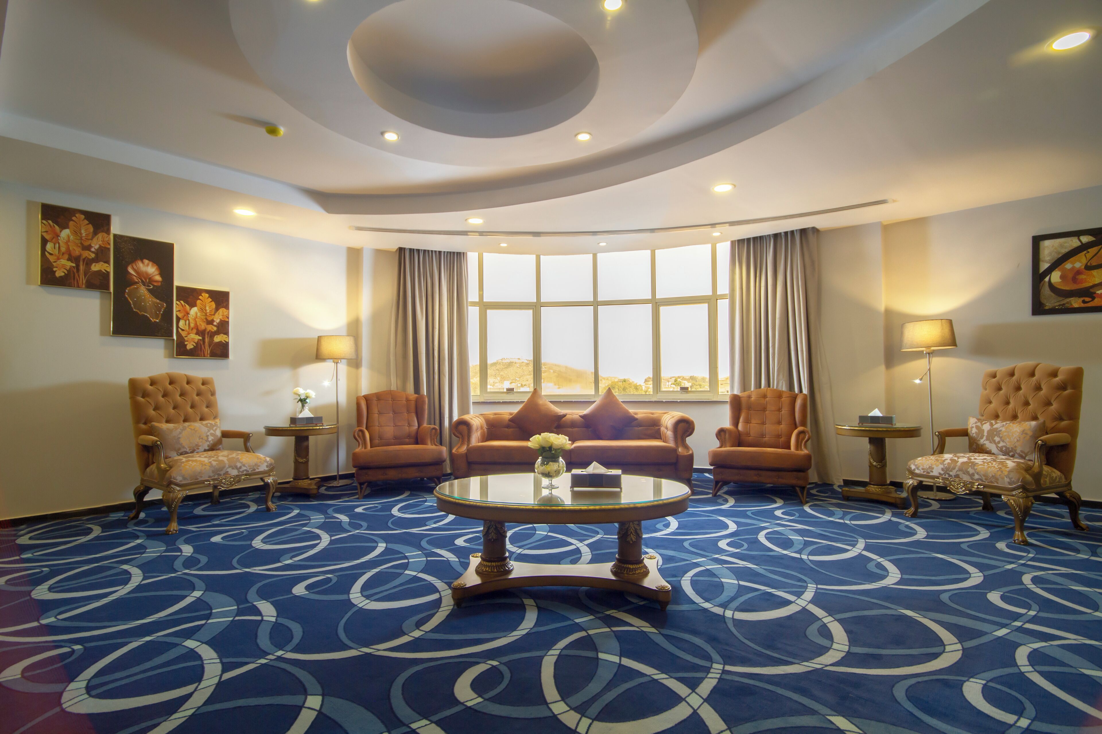 A hotel room with a round coffee table, two armchairs, a sofa, a large window with curtains, and a patterned carpet.