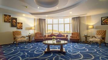 A hotel room with a round coffee table, two armchairs, a sofa, a large window with curtains, and a patterned carpet.