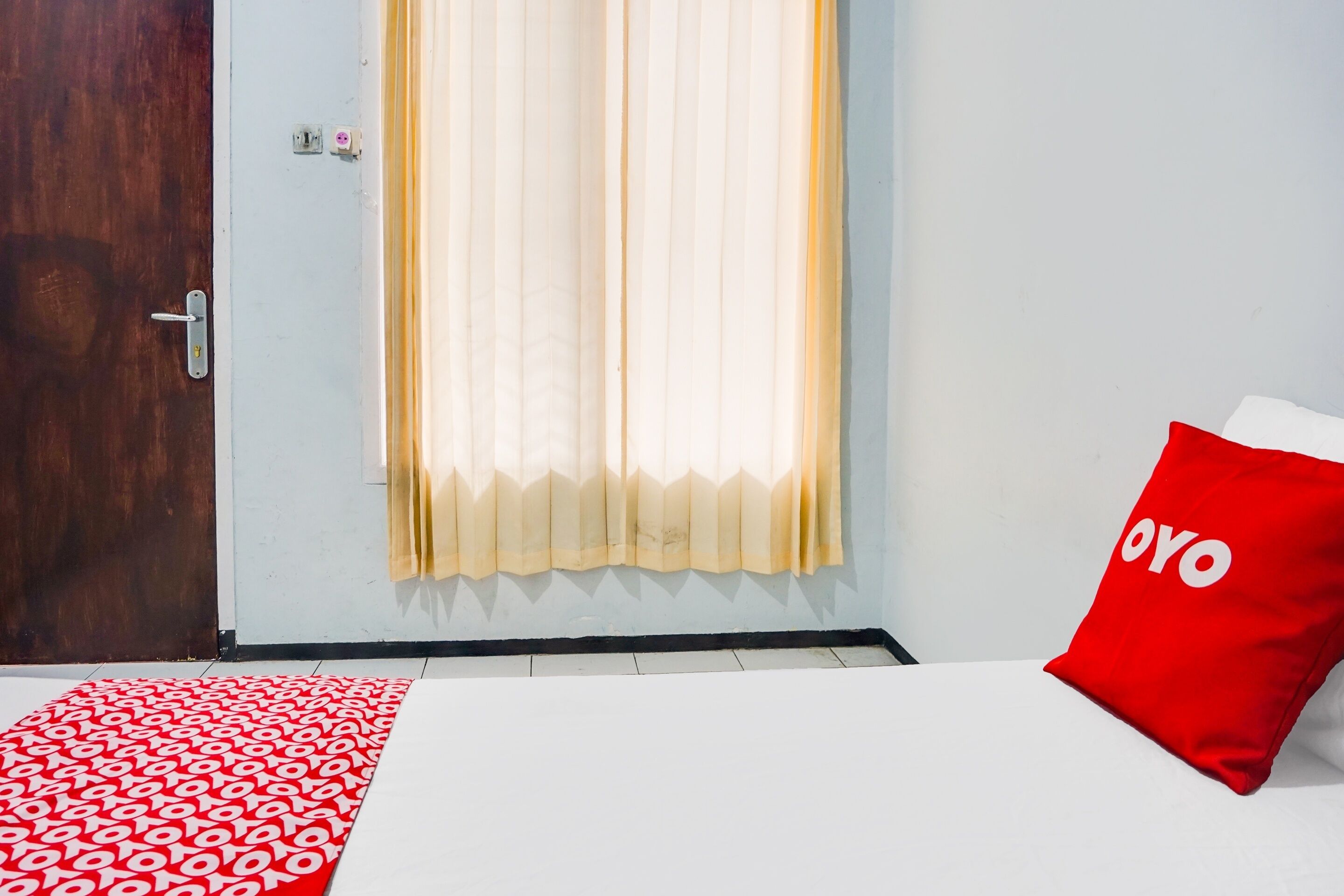 Una habitación con una cama, un almohadón rojo con el texto 'OYO', un tapete rojo y blanco estampado y una ventana con cortinas de encaje.