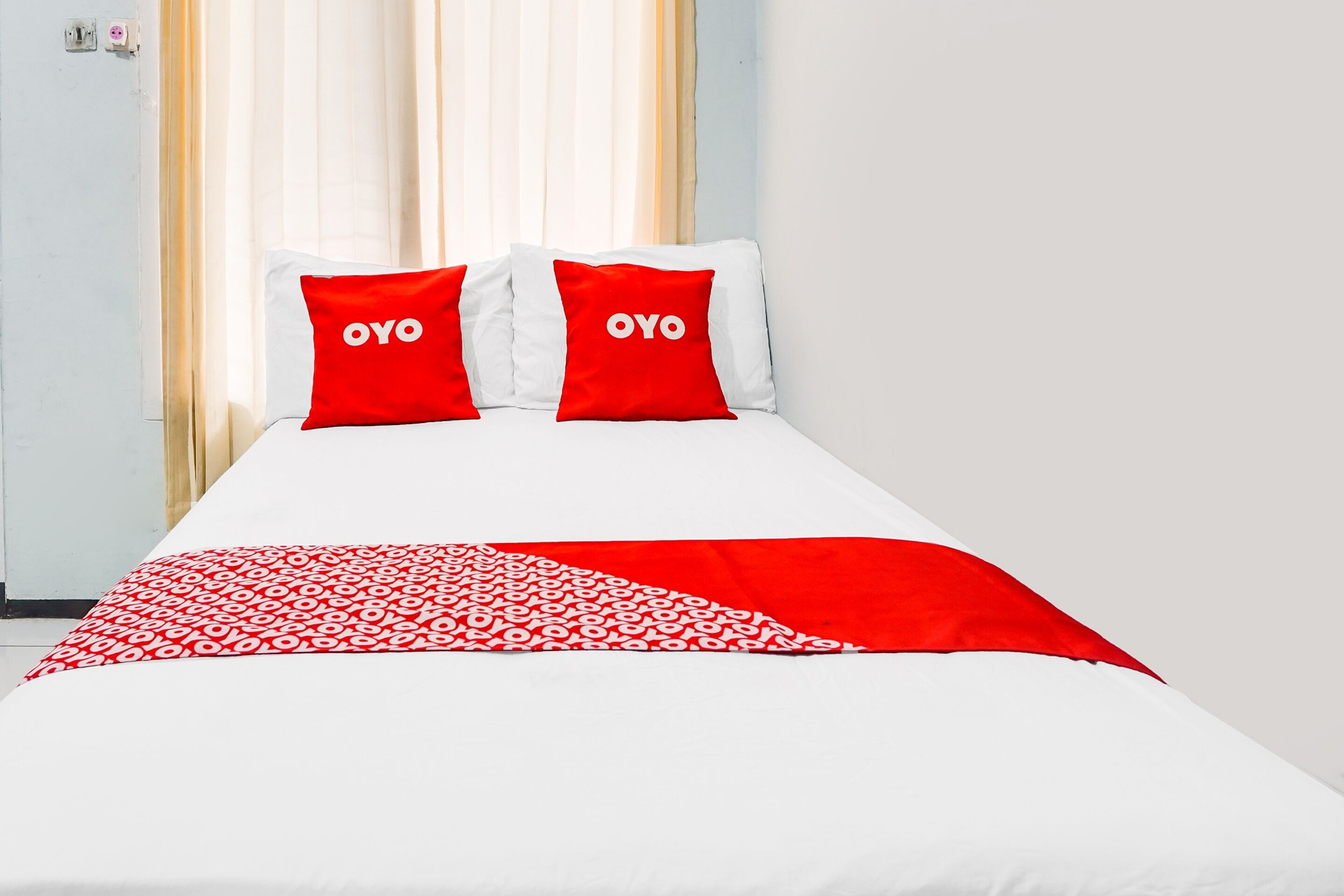 Habitación de hotel con cama, dos almohadas con la marca "OYO", una manta estampada en rojo y blanco, y una ventana con cortinas.