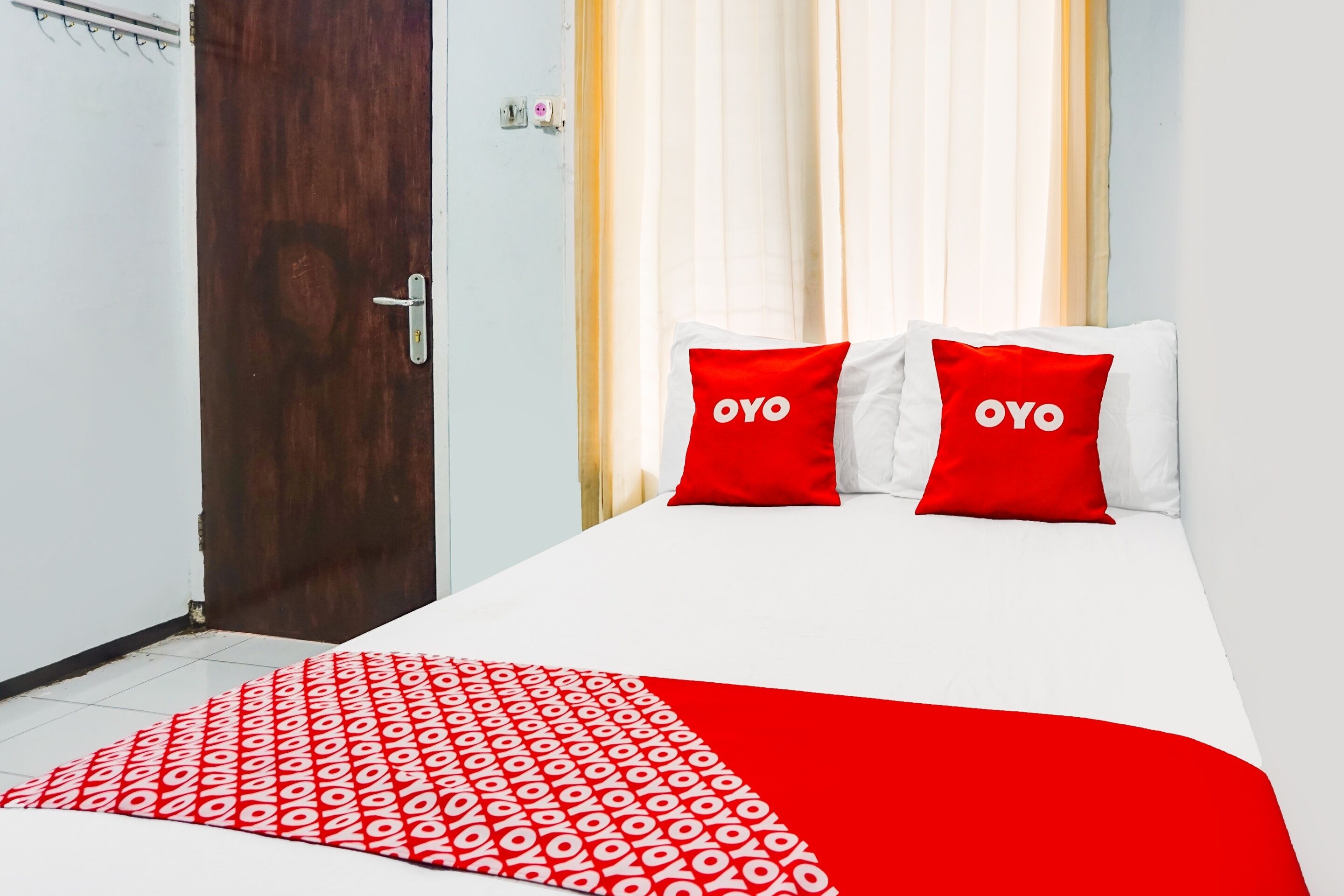 Et hotelværelse med en seng, to røde puder med teksten "OYO", et rødt og hvidt mønstret tæppe og en trædør.