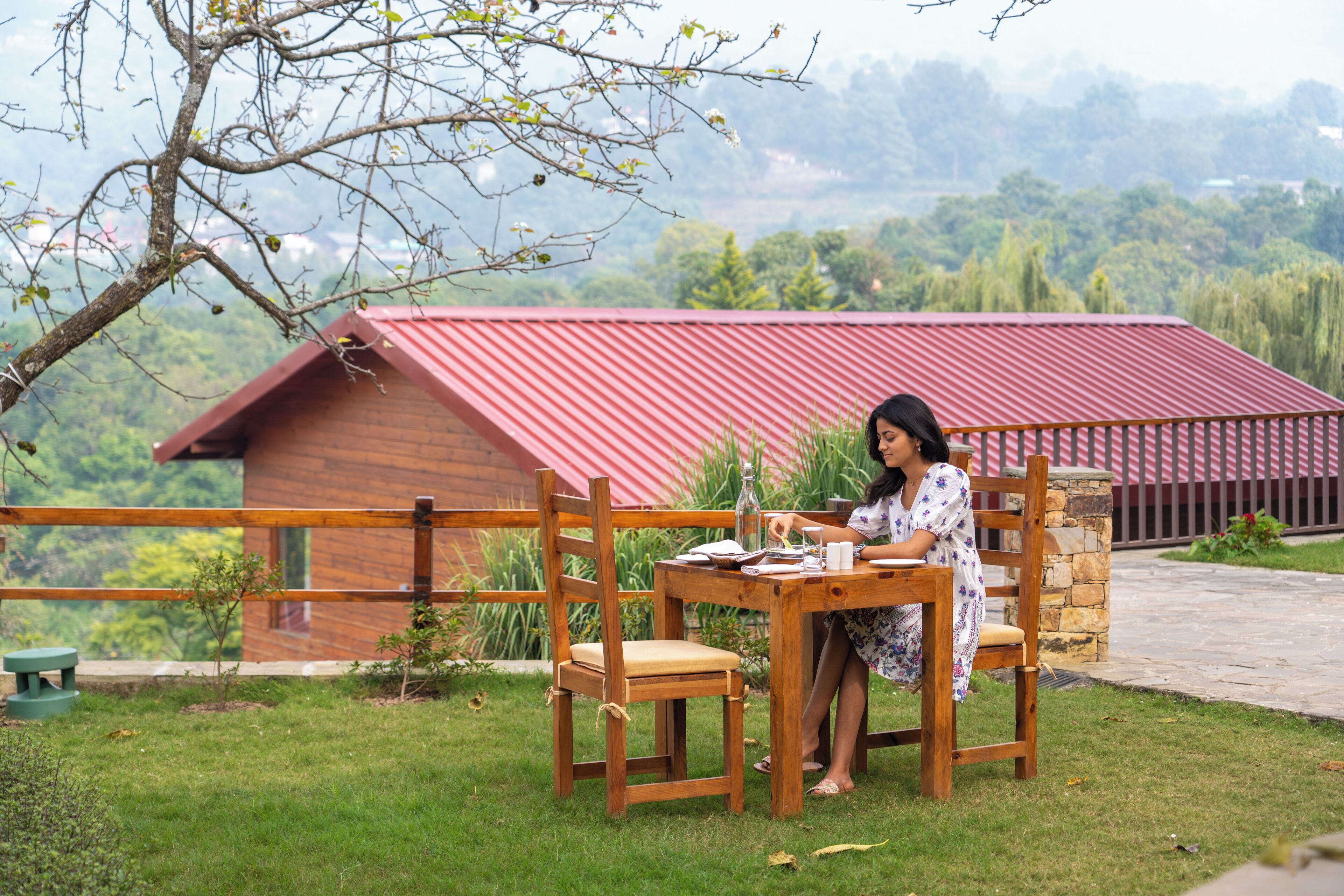 Une femme est assise à une table en bois, avec un bâtiment à toit rouge en arrière-plan.
