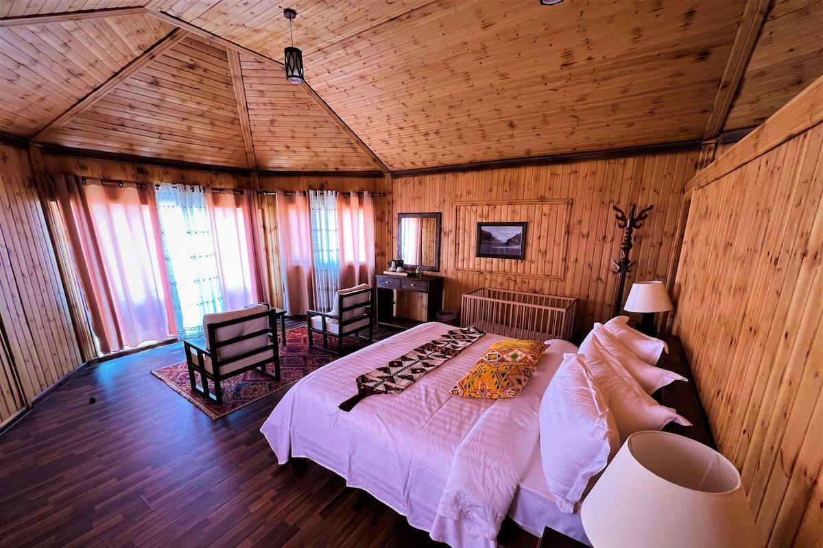 A wooden cabin bedroom with a bed, two chairs, a desk, and a crib.
