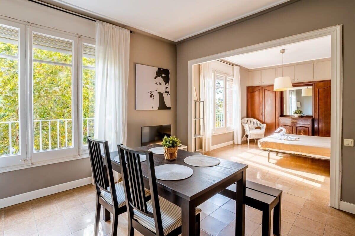 Un comedor con mesa y sillas, un cuadro en la pared y vista a un dormitorio con cama y sillón.