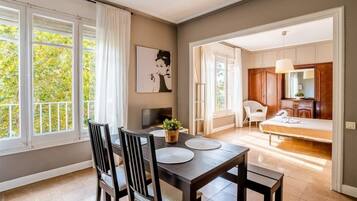 Un comedor con mesa y sillas, un cuadro en la pared y vista a un dormitorio con cama y sillón.