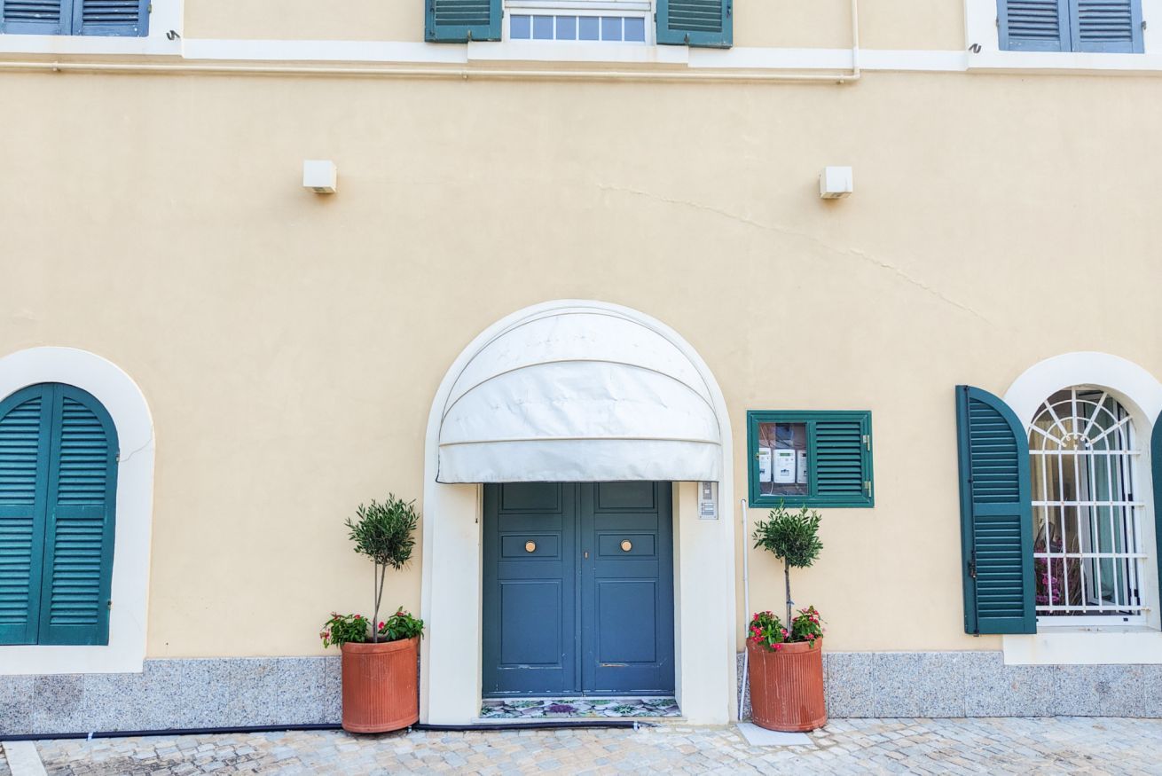 Fachada de edificio con una puerta azul central, flanqueada por dos macetas y un toldo blanco.