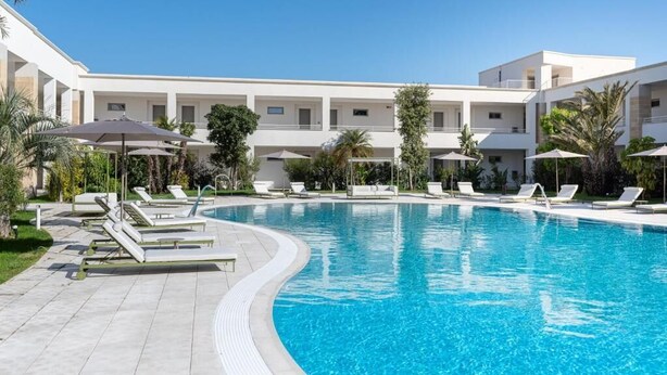 Una piscina con sillas de descanso y sombrillas, rodeada por un edificio de varios pisos con balcones.