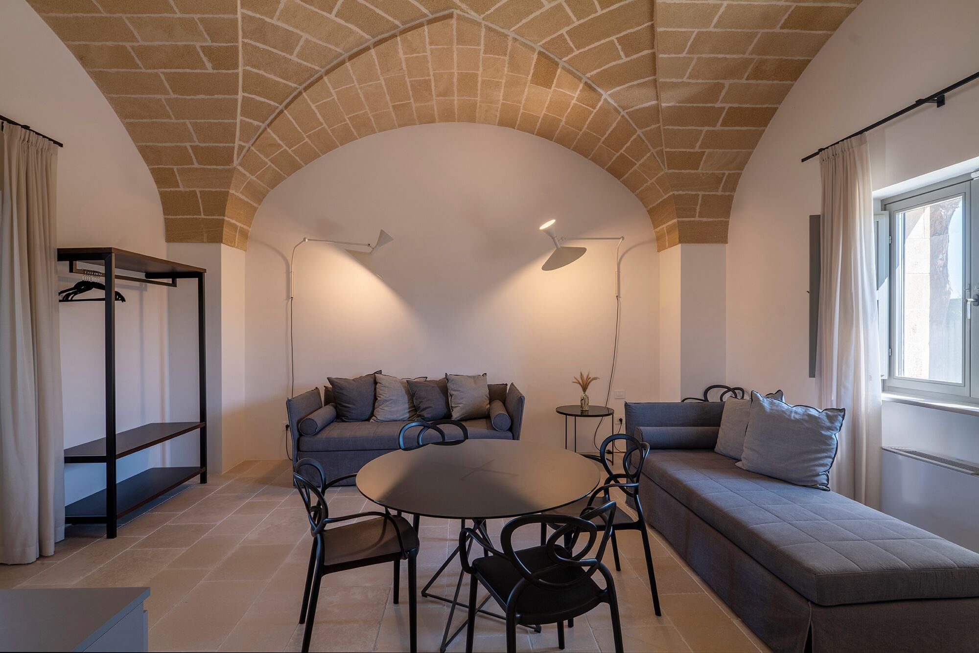Un salon chaleureux avec un canapé, une table ronde et deux chaises. On y trouve un luminaire suspendu et une fenêtre avec des rideaux.