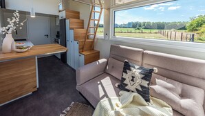 En kompakt opholdsstue med et tekøkken, en sofa med et pled og en stige til et hvileloft, samt et stort vindue med udsigt over grøn bevoksning.
