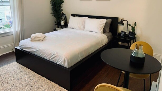 A modern bedroom with a bed, bedside table, lamp, and a small round table with a vase. - Pet-Friendly, Lovely Studio w/ Balcony (Halifax)