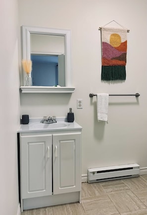 A bathroom with a white vanity, a mirror, a towel rack with a white towel, a colorful tapestry, and a white heater on the floor.