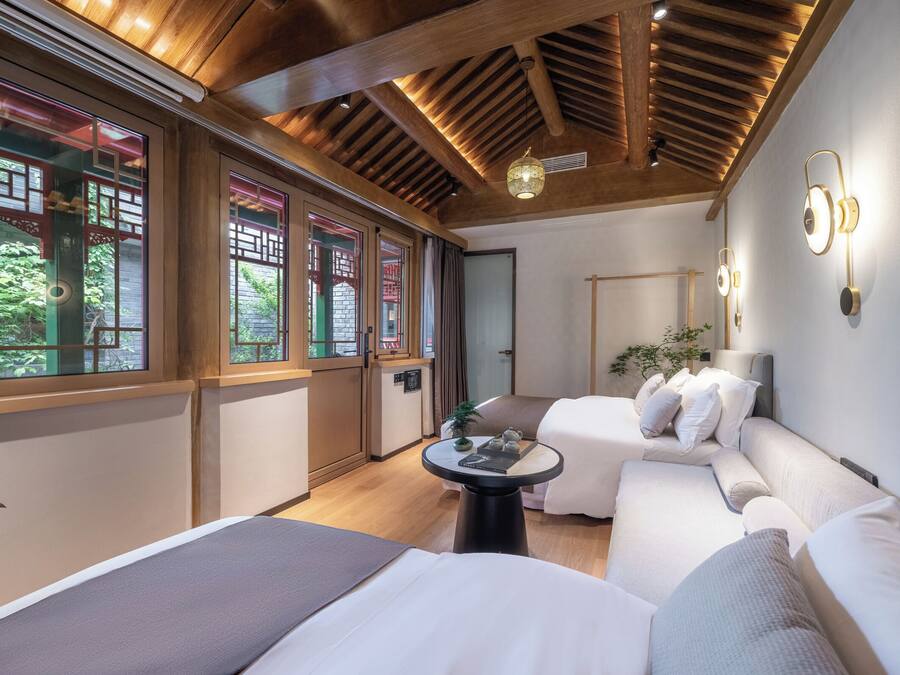 Una habitación de hotel moderna con vigas de madera en el techo, ventanas grandes con diseño de celosía tradicional, un sofá cama, una pequeña mesa redonda e iluminación empotrada.