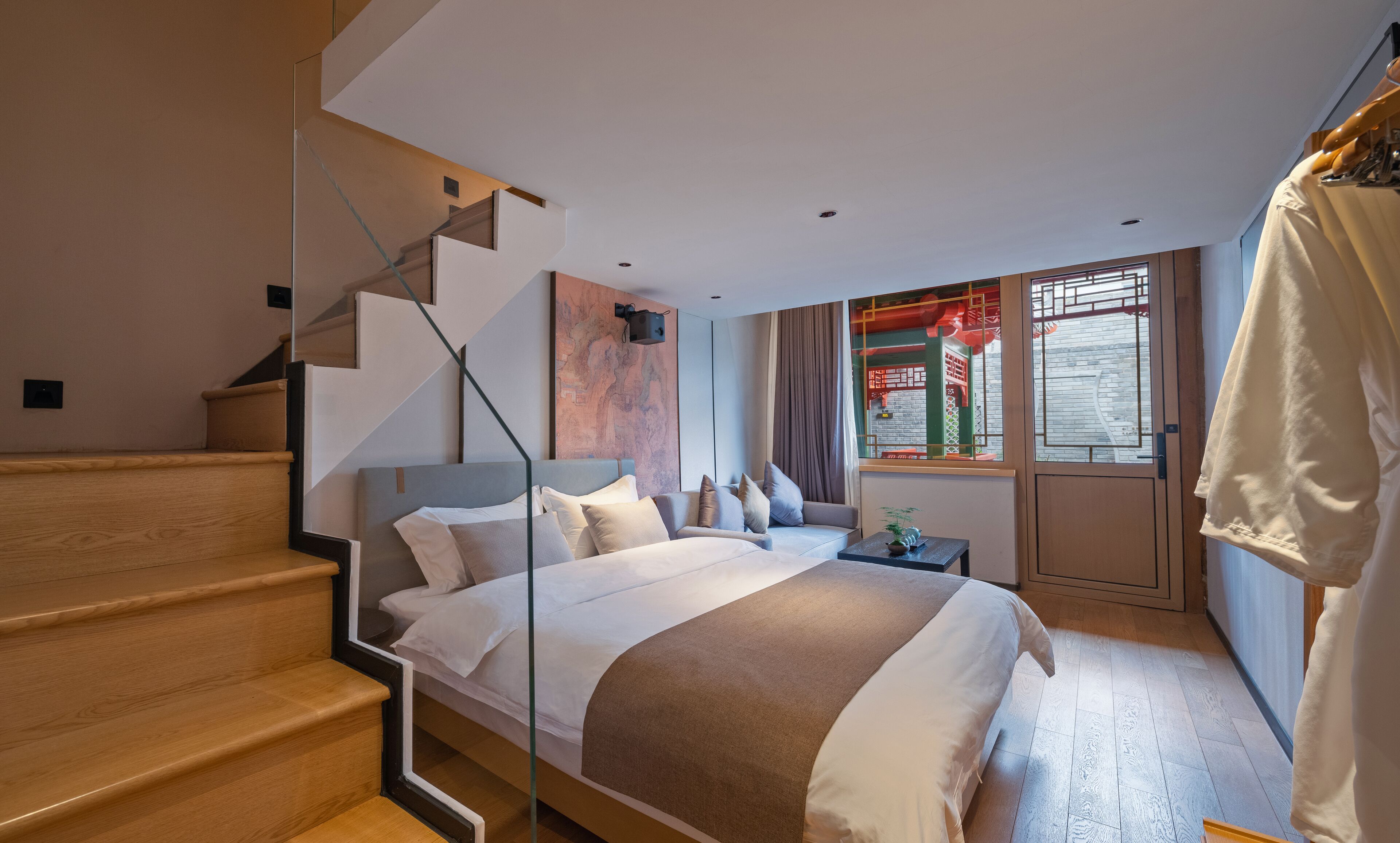 A modern bedroom with a large bed, a glass staircase, a small table with a plant, and a view of a traditional building through a window.