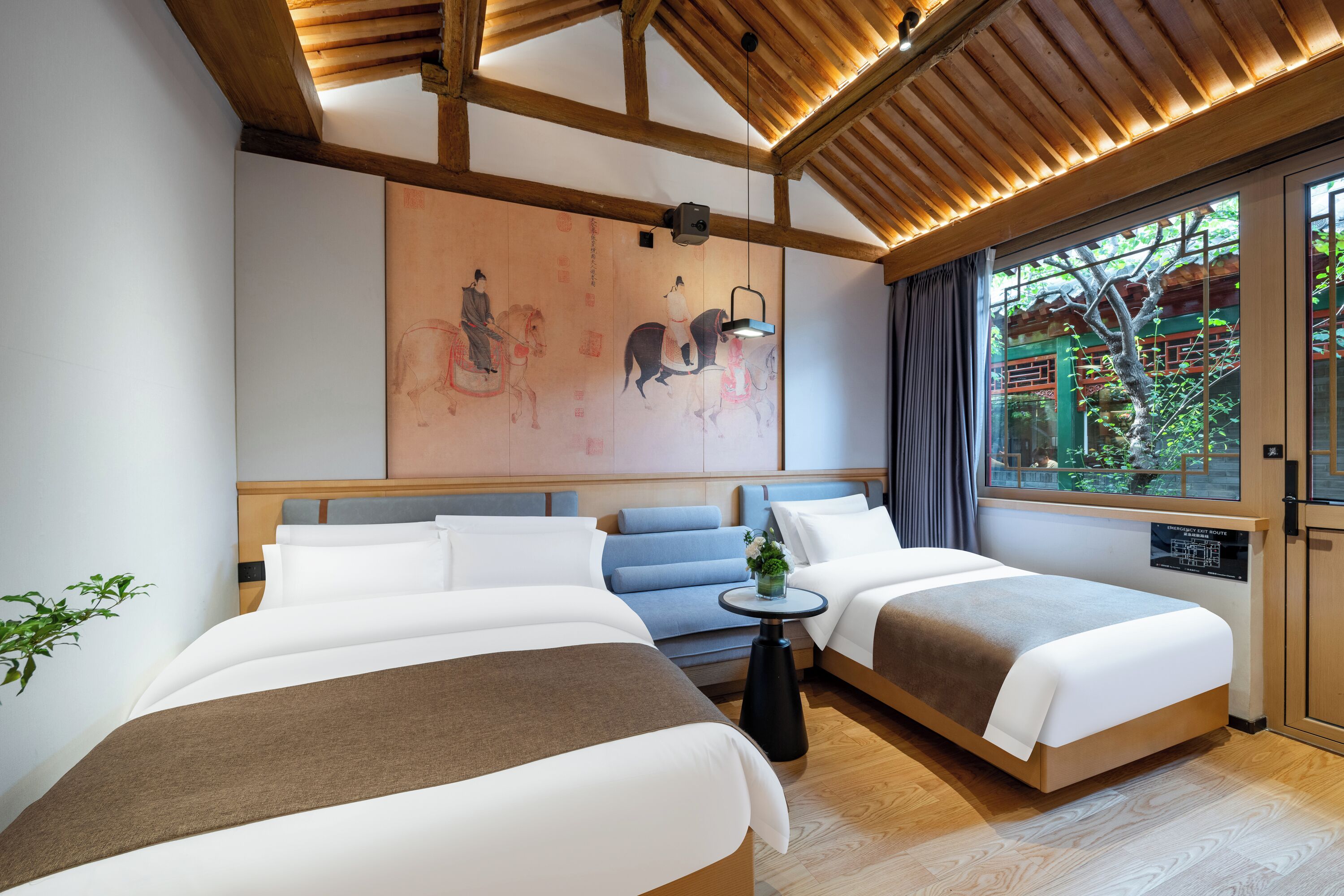 A hotel room with two beds, a wooden ceiling, a large window with a view of greenery, and a traditional painting on the wall.