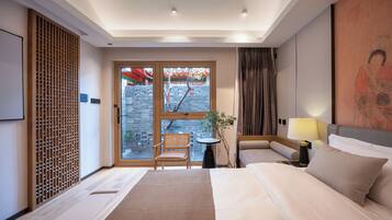 Ein Hotelzimmer mit einem Bett, zwei Nachttischen, einem Sessel, einem Fenster mit Blick auf eine Ziegelwand und Bäume sowie einer Wandverkleidung.